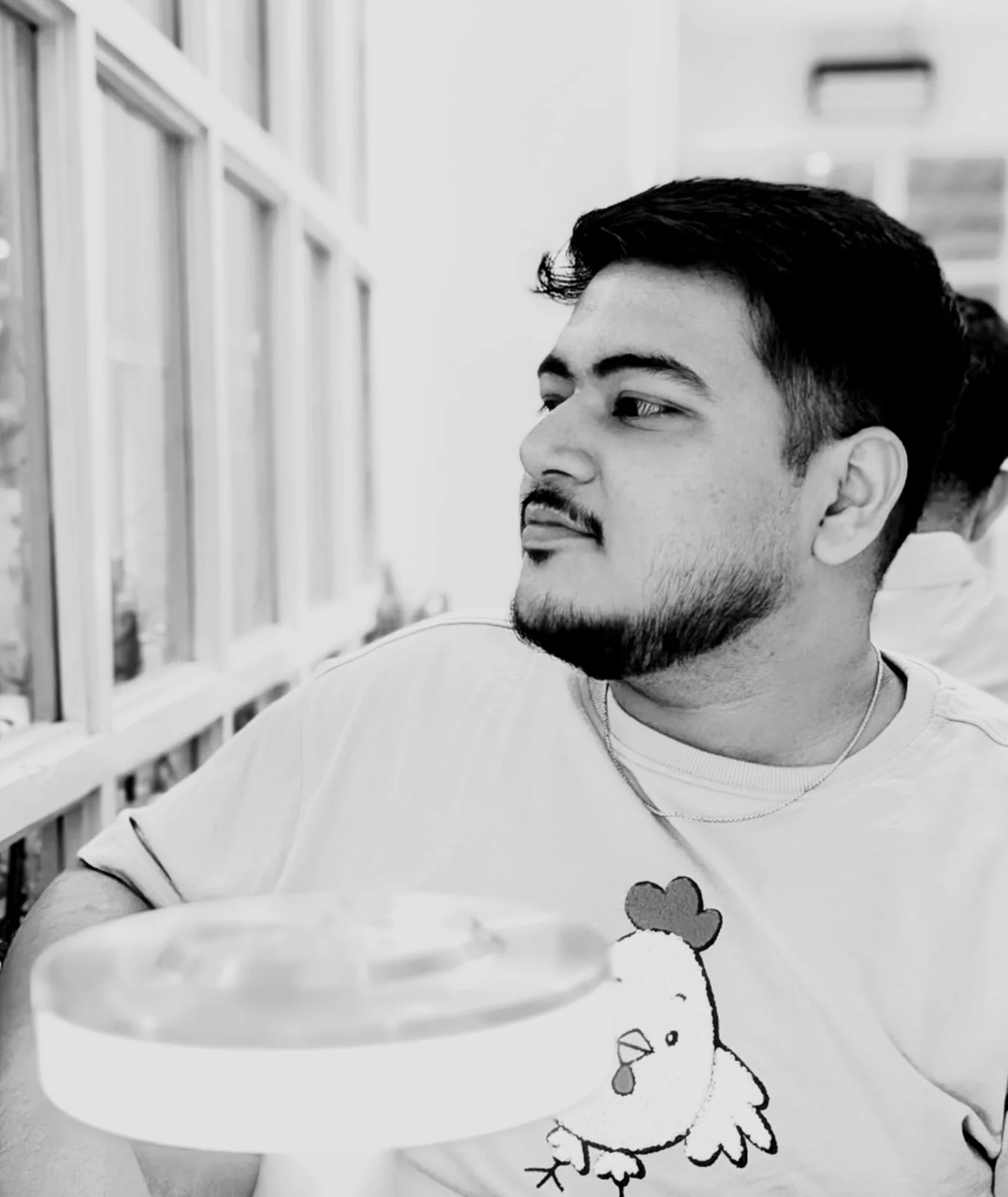 Praduan Saha in black and white, seated beside a row of tall windows and looking out of them